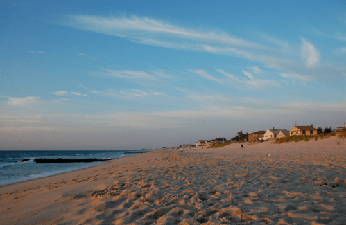 A serene beach scene with soft sand, gentle waves, and houses lining the shore under a blue sky with wispy clouds.