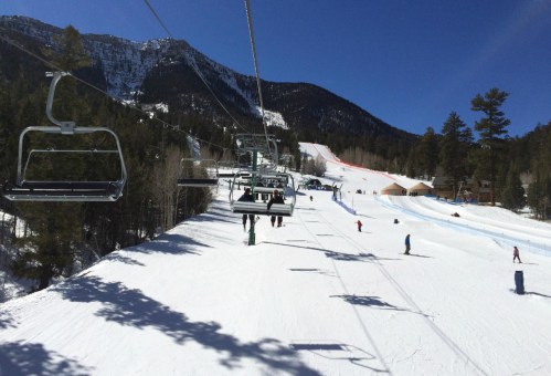 A ski lift ascends a snowy mountain with skiers on the slopes below and trees lining the path.