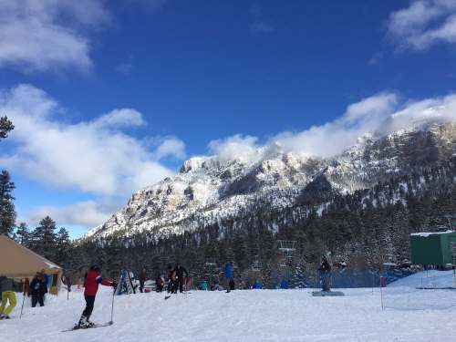 A snowy landscape with skiers, mountains in the background, and a clear blue sky with scattered clouds.