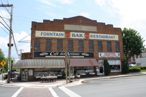 Historic brick building with signs for a bar and restaurant, featuring outdoor seating and nearby trees.