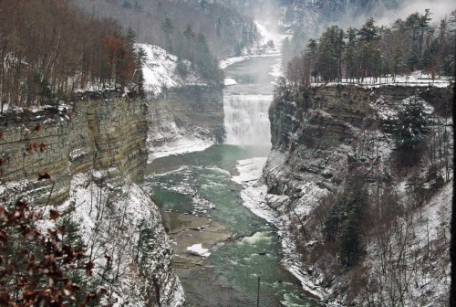 A scenic view of a river flowing through a snowy canyon, with a waterfall in the distance and mist rising from the water.