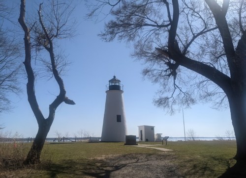 A white lighthouse stands near a calm waterway, surrounded by bare trees under a clear blue sky.
