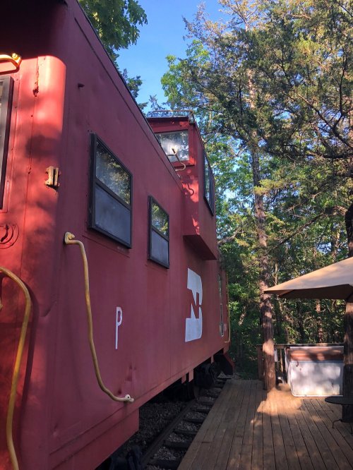 A red train caboose nestled among trees, with a wooden deck and a hot tub visible nearby.