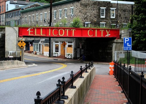 Red bridge with "Ellicott City" sign over a road, surrounded by stone buildings and greenery.