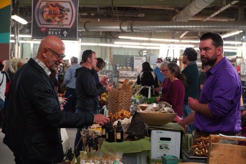 A bustling market scene with people browsing stalls, featuring mushrooms, produce, and various goods for sale.