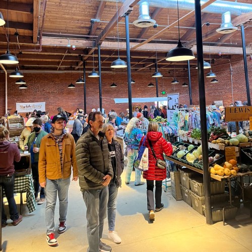 A bustling indoor market with people browsing stalls filled with colorful clothing and fresh produce.