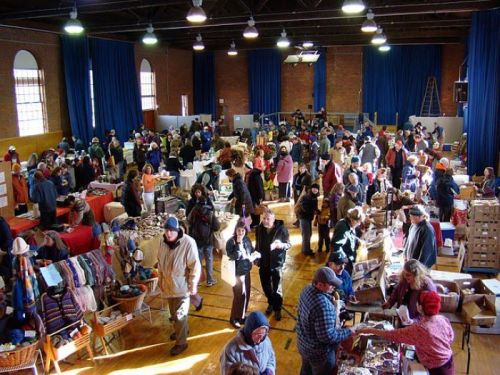 A bustling indoor market with many vendors and shoppers browsing various handmade goods and crafts.