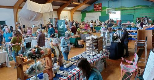 A bustling indoor market with vendors and shoppers, showcasing various goods and crafts in a community hall.