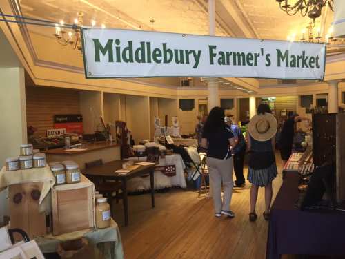 A bustling farmer's market with a banner reading "Middlebury Farmer's Market" and various vendor displays.