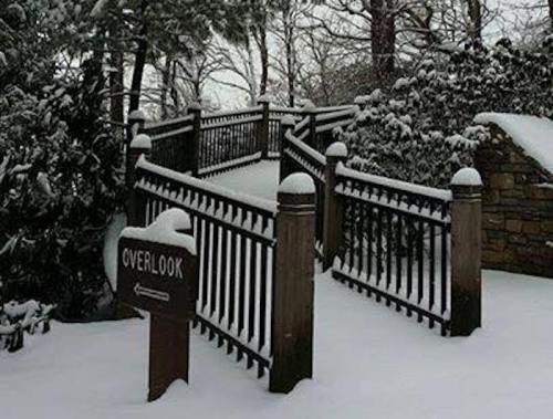 A snow-covered overlook path with a wooden railing and a sign pointing towards the viewpoint. Trees in the background.