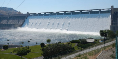 A large dam releasing water, creating a cascading waterfall effect, with greenery and pathways in the foreground.