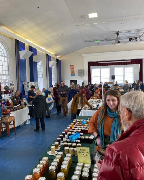 A bustling indoor market with vendors and shoppers, showcasing jars of pickles and various local products.