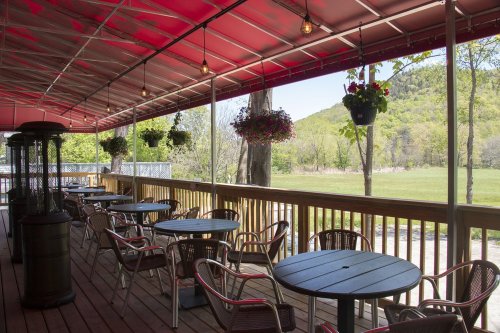 A cozy outdoor patio with tables and chairs, surrounded by greenery and hanging flower baskets under a red canopy.
