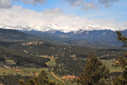 A panoramic view of snow-capped mountains over a lush green valley, dotted with trees and a winding road.