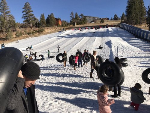 People carrying inner tubes on a snowy slope, with others tubing down the hill in a winter recreation area.