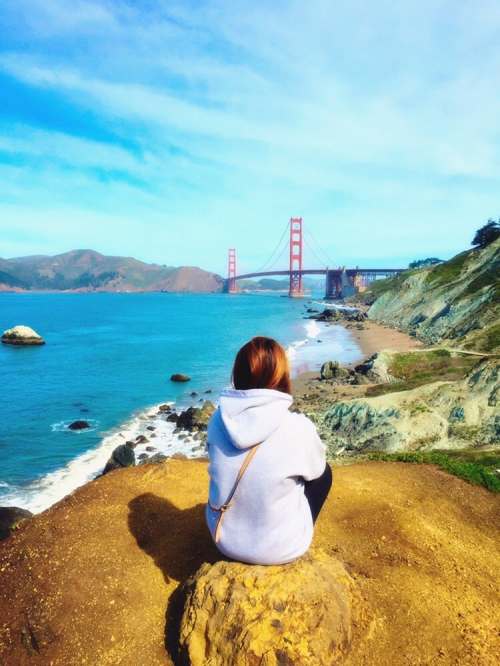 A person sits on a rock by the shore, overlooking the Golden Gate Bridge and the ocean under a clear blue sky.