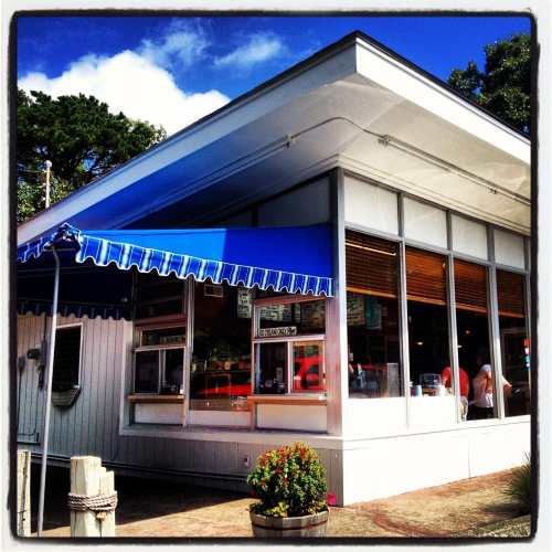 A bright, modern eatery with a blue awning and large windows, surrounded by greenery and a clear blue sky.
