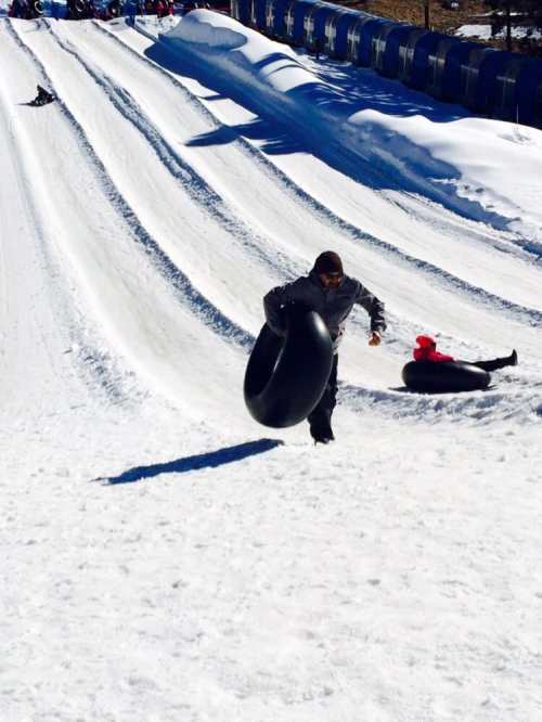 A person pulls a black inner tube up a snowy hill, with others tubing in the background.