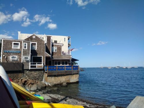 A seaside building with a wooden exterior, overlooking calm waters and boats in the distance under a clear blue sky.