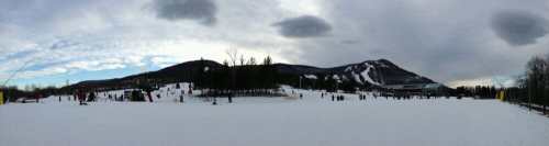 A panoramic view of a snowy landscape with a ski resort and mountains under a cloudy sky.