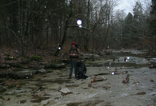 A person stands by a rocky stream in a forest, wearing a red hat, with snowflakes falling around them.