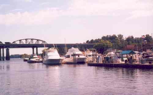 A serene marina with several boats docked, under a bridge and surrounded by trees on a sunny day.