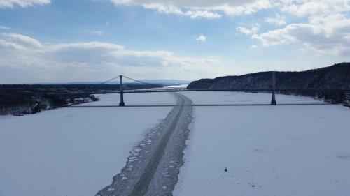 Aerial view of a frozen river with a bridge spanning across, surrounded by snow-covered hills and a cloudy sky.