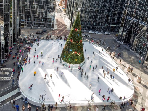A bustling ice skating rink surrounded by skyscrapers, featuring a large decorated Christmas tree at the center.