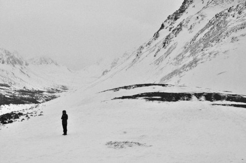 A lone figure stands on a snowy landscape, surrounded by mountains under a cloudy sky.