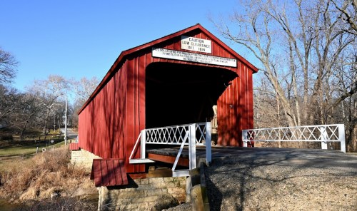 A red covered bridge with a low clearance sign, surrounded by bare trees and a gravel path.