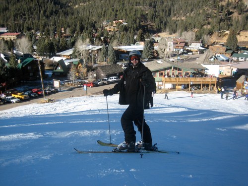 A skier in black gear stands on a snowy slope with a mountain town in the background.