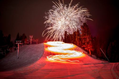 Fireworks illuminate a snowy slope at night, with trails of light from skiers creating a vibrant scene.