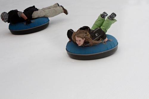 Two people sledding on blue inflatable tubes down a snowy slope, one lying face down and the other in a seated position.