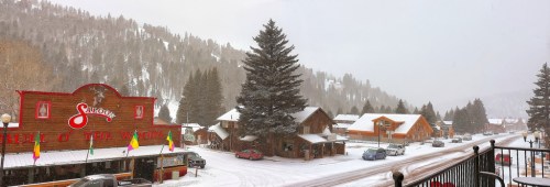 Snowy street scene in a small town, featuring wooden buildings and a large sign, with trees and mountains in the background.