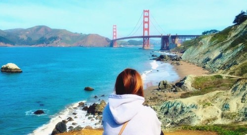 A person sits on a rocky shore, overlooking the ocean and the Golden Gate Bridge in the distance.