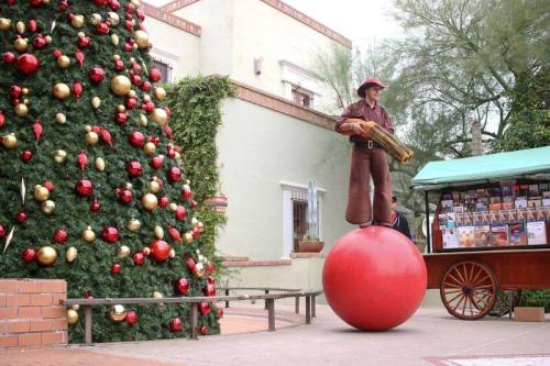 A performer stands on a large red ball next to a decorated Christmas tree and a cart filled with books.