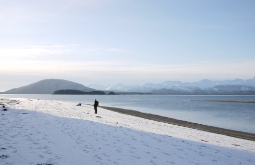 A person stands on a snowy beach by a calm lake, with mountains in the background under a clear sky.