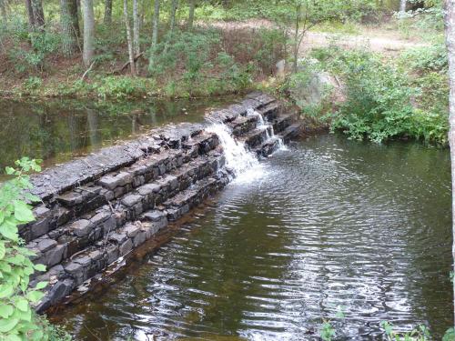 A small waterfall cascades over a stone structure into a calm pond, surrounded by lush greenery and trees.
