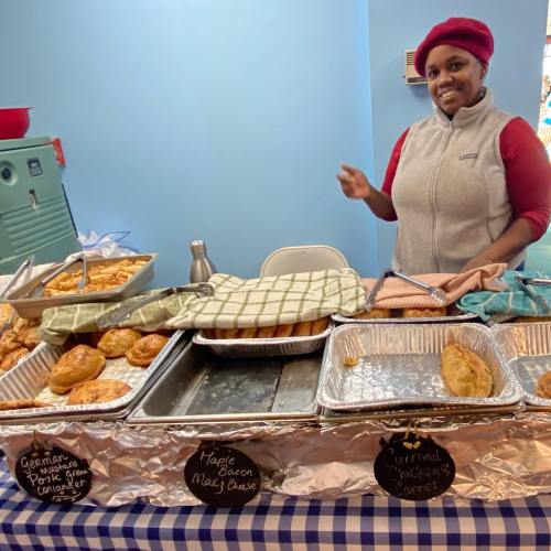 A smiling vendor stands behind a table with various baked goods, including pastries and savory items, in a bright setting.