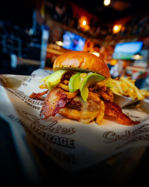 A close-up of a burger with bacon, avocado, and crispy onions, served with fries in a casual dining setting.