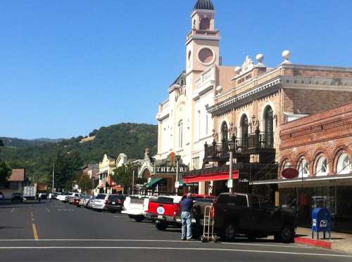 A sunny street scene with parked cars, historic buildings, and a theater in a small town. Green hills in the background.