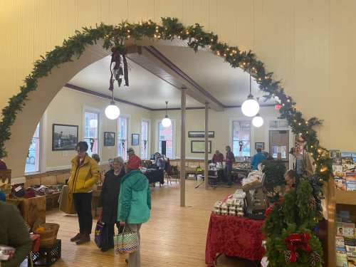 A festive market scene with people shopping under a decorated arch, featuring holiday decorations and various stalls.