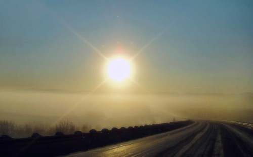A bright sun rises over a foggy landscape, illuminating a winding road in the foreground.