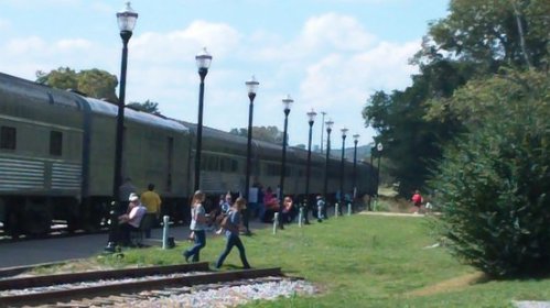 A train parked beside a grassy area with people walking along the tracks and lamp posts lining the path.