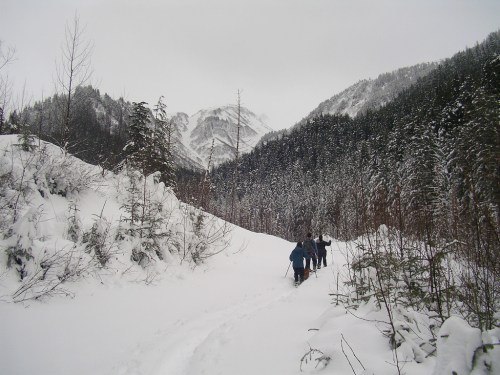 Two hikers trek through a snowy landscape, surrounded by mountains and evergreen trees under a cloudy sky.