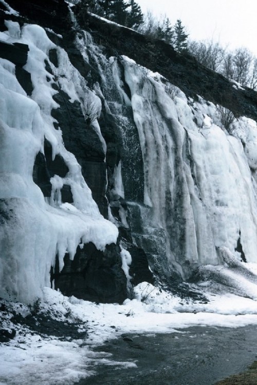 A frozen waterfall cascading down a rocky cliff, surrounded by snow and ice, with a small stream at the base.