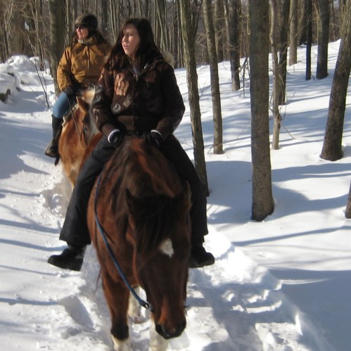 Two people riding horses through a snowy forest, surrounded by tall trees.