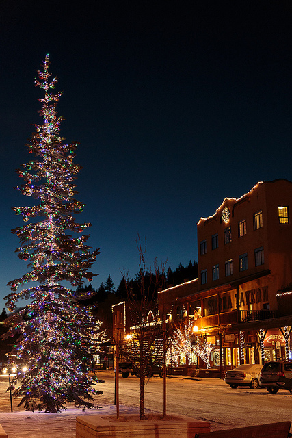A brightly lit Christmas tree stands in a snowy town square, with festive lights adorning nearby buildings at dusk.