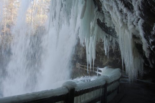 Icicles hang from a rocky overhang near a frozen waterfall, with a wooden railing in the foreground.