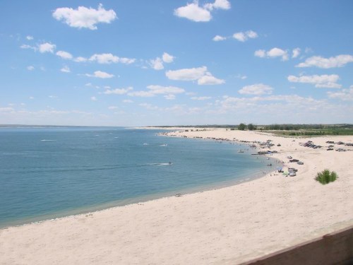 A sandy beach with clear blue water, dotted with boats and cars, under a bright sky with fluffy clouds.